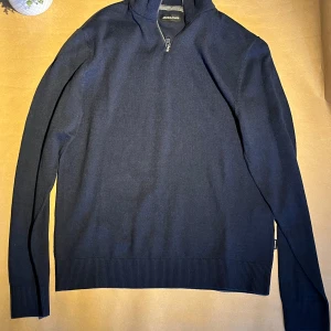 Mörkblå stickad tröja med dragkedja från Jack & Jones - Mörkblå stickad tröja från Jack & Jones med halv dragkedja vid halsen. Tröjan har lång ärm och ribbade muddar vid ärmslut och nederkant. Perfekt för lager-på-lager och enkel att matcha.