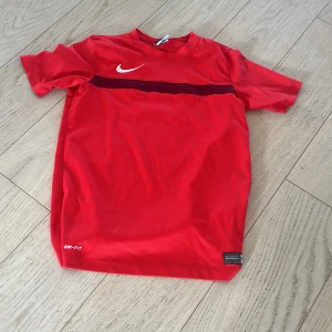 Röd sportig t-shirt från Nike - Röd t-shirt från Nike med vit logga på bröstet och en mörkröd rand över bröstet. Tillverkad i ett lätt och ventilerande material, perfekt för träning eller sport. Klassisk passform med korta ärmar och rund hals.