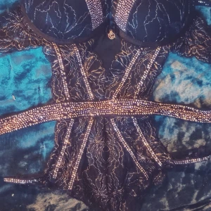 Svart glittrig body med spetsdetaljer - Säljer en svart body med glittriga pärldetaljer och vackert spetsmönster. Bodyn har vadderade kupor och markerad midja, perfekt för att skapa en snygg siluett. Snygga band och genomarbetade detaljer ger en het sommar look.