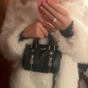 Svart handväska från Zadig & Voltaire - Säljer en svart handväska från Zadig & Voltaire i skinn med silverfärgade dragkedjor och detaljer. Väskan har två handtag, avtagbar axelrem med logga och dekorativa nitar. Perfekt accessoar för att lyfta din outfit. Dustbag tillkommer