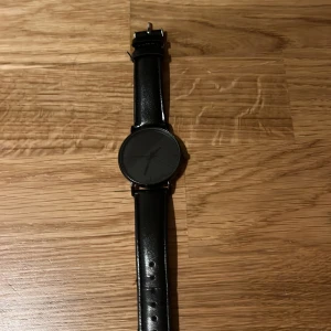 Svart minimalistisk klocka  - Stilren svart klocka med svart urtavla och svarta visare. Klockan har ett enkelt, modernt utseende och svart läderarmband med slät finish. Perfekt för dig som gillar en clean look och diskreta accessoarer.