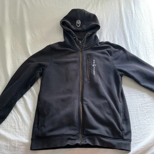 Svart hoodiejacka från Sail Racing - Svart hoodiejacka från Sail Racing med dragkedja framtill och på bröstfickan. Jackan har broderad logga på huvan och bröstet samt mjukt fleecefoder på insidan. Perfekt för kyliga dagar. Obs lite slitna vid armbågarna.