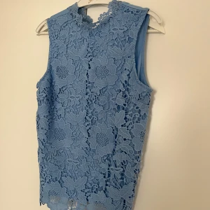 Blå spetsblus med blommönster - Säljer en ljusblå ärmlös blus med vackert blommigt spetsmönster och hög hals. Blusen har en mjuk underklänning och knäppning i nacken. Perfekt för dig som vill ha en feminin och stilren topp.