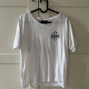 Vit t-shirt från Dope med tryck - Vit t-shirt från Dope med svart tryck framtill och stort tryck på ryggen med texten 'DOPE Paradise isn't tropical' och bergmotiv. Rund halsringning och normal passform. Perfekt för en avslappnad stil.