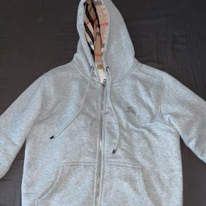 Grå hoodie med dragkedja från Burberry - Säljer en grå hoodie från Burberry med klassiskt rutigt foder i huvan och insidan. Tröjan har dragkedja framtill, två fickor och justerbar huva med snören. Perfekt för en stilren och bekväm look.
