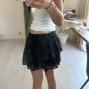 Svart volangkjol - Säljer en svart kortkjol med volanger och diskret prickigt mönster. Kjolen har resår i midjan och är tillverkad i ett lätt, transparent tyg. Perfekt för dig som gillar en luftig och trendig stil.perfekt nu till sommaren