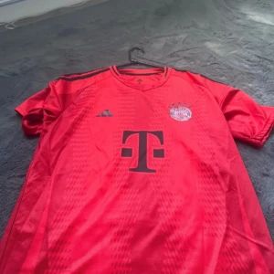 Bayern München fotbollströja med Kane 9 - Röd Bayern München matchtröja från Adidas med svart tryck, klubbmärke och sponsorlogga på bröstet. Nummer 9 och namnet Kane på ryggen. Tröjan har korta ärmar och är tillverkad i ett lätt funktionsmaterial.