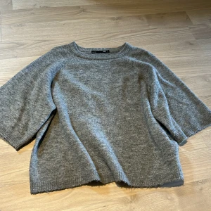 Grå stickad tröja från 157 - Mysig grå stickad tröja från 157 med rund halsringning och kortare ärmar. Perfekt för lager-på-lager och enkel att matcha med olika outfits. Klassisk och stilren design.