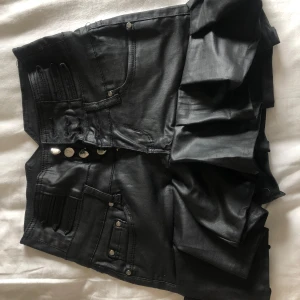 Svart volangkjol  - Snygg svart kjol i jeansmaterial med volangdetalj nedtill. Kjolen har klassiska jeansfickor fram och bak samt knappar framtill. Perfekt för dig som vill ha en edgy och cool stil.