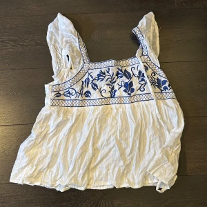 Vit blus med blå broderier - Säljer en luftig vit blus med breda axelband och vackra blå broderade blommor framtill. Blusen har band på ryggen som går att knyta till en rosett. Oanvänd då den var för liten vid bysten 