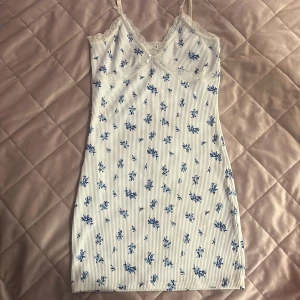 Vit blommig spetsklänning från H&M Divided - Söt vit klänning med blå blommor från H&M Divided. Klänningen har smala axelband, spetsdetaljer vid bysten och en liten rosett framtill. Den är kort och har en figurnära passform, perfekt för varma dagar.