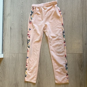 Ljusrosa leggings med blommiga revärer - Säljer ett par ljusrosa leggings med elastisk midja och blommiga revärer längs båda sidorna. Perfekta för en färgglad och bekväm stil. Mjukt material och smal passform.