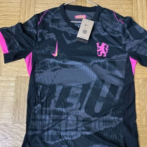 Chelsea tröja third kit  - Säljer en svart fotbollströja från Nike med rosa och gula detaljer. Tröjan har ett mönster över hela tyget, rosa lejonemblem på bröstet och gula inslag vid sidorna. Kortärmad modell med sportig känsla och modern design.