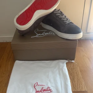 Gråa sneakers från Christian Louboutin med röd sula - Säljer ett par gråa sneakers från Christian Louboutin med den ikoniska röda sulan. Skorna har snörning, vit platt sula och är tillverkade i mocka.  Passar till de flesta använda Max 5-6 gånger 