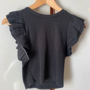 Svart volangtopp från Zara - Svart ribbad topp från Zara med volangdetaljer på axlarna. Toppen har rund halsringning och är ärmlös med markerade volanger som ger en trendig look.
