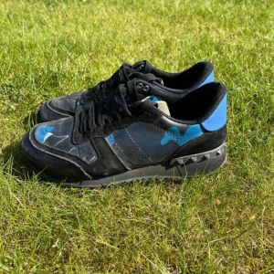 Valentino Rockrunner sneakers med blå kamouflage - Säljer ett par Valentino Rockrunner sneakers med svart och blå kamouflage, svarta snören och detaljer i mocka och skinn. Skorna har den klassiska nitarna på sulan och en cool, sportig look. Perfekta för dig som gillar unika och trendiga sneakers.
