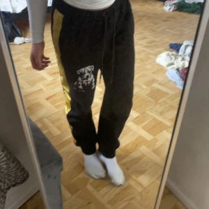 Svarta mjukisbyxor från Amstaff - Svarta mjukisbyxor med gul sidopanel och vitt tryck på ena benet. Byxorna har resår i midjan och dragsko för justerbar passform. Perfekta för en avslappnad stil och sköna att ha på sig hemma eller till träning.