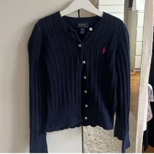 Mörkblå kabelstickad kofta från Polo Ralph Lauren - Säljer en klassisk mörkblå kabelstickad kofta från Polo Ralph Lauren med knappar framtill och broderad rosa logga på bröstet. Tröjan har långa ärmar och ribbade muddar. Perfekt för lager-på-lager-stil.