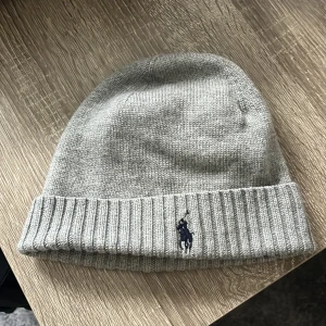 Grå stickad mössa från Polo Ralph Lauren - Snygg grå stickad mössa från Polo Ralph Lauren med klassisk ribbad kant och broderad logga framtill. Perfekt accessoar för kyliga dagar och enkel att matcha med olika outfits.