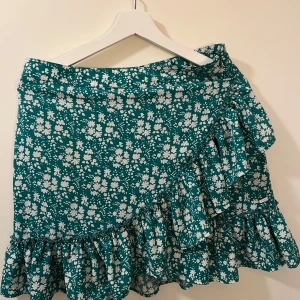 Grön blommig omlottkjol från Ghanda - Säljer en grön omlottkjol från Ghanda med vitt blommönster. Kjolen har volangdetaljer längs nederkanten och stängs med dragkedja i sidan.