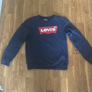 Mörkblå sweatshirt från Levi's - Säljer en mörkblå sweatshirt från Levi's med klassiskt rött och vitt logotryck på bröstet. Tröjan har rund halsringning och långa ärmar. Perfekt för en avslappnad och stilren look.