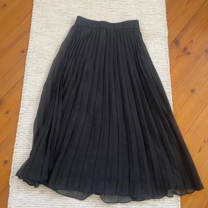 Svart plisserad långkjol från Monki - Säljer en svart plisserad långkjol från Monki. Kjolen har resår i midjan och är tillverkad i ett lätt, flödande material som ger ett snyggt fall. Perfekt att styla med sneakers eller boots för en cool look.