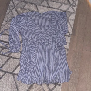 Blåvit omlottklänning med ränder - Säljer en blåvit omlottklänning med smala vertikala ränder och trekvartsärm. Klänningen har knytband i midjan och en luftig passform, perfekt för varmare dagar.