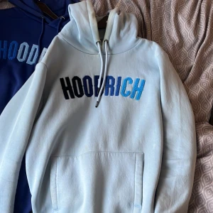 Ljusblå hoodie från Hoodrich - Säljer en ljusblå hoodie från Hoodrich med broderad logga i svart, mörkblå och blått framtill. Tröjan har huva med dragsko och en stor magficka. Perfekt för dig som gillar streetwear och vill ha en snygg och bekväm tröja.