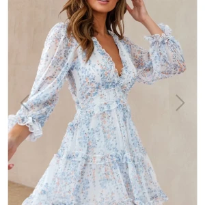 Blommig volangklänning med öppen rygg, stl XS - Superfin kortklänning i vitt med blått och ljusrosa blommönster från Dennis Maglic, använd endast en gång! Klänningen har volangdetaljer, v-ringning och öppen rygg. Långa, transparenta ärmar med resår vid ärmslut. Perfekt för vår och sommar, eller varför inte till Midsommar🌸! Nypris 899kr.