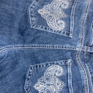 Blå jeans med broderade bakfickor från Lee - Säljer ett par blå jeans från Lee med snygga broderade detaljer på bakfickorna. Jeansen har låg midja och klassisk femficksdesign. Perfekta för dig som vill ha ett par jeans med lite extra stil.