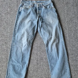 Levis 501 ljusblå jeans - Klassiska Levis 501 jeans i ljusblå denim med raka ben och femficksmodell. Byxorna har knappgylf och slitna detaljer vid benslut och ett hål på låret för en extra vintagekänsla. Perfekta för dig som gillar en avslappnad och tidlös stil. Storlek 30x32