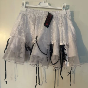 Kjol fr Poizen onesize - Supersöt vit kjol i spets med svarta kedjor, korsdetalj och rosetter. Kjolen har elastisk midja och flera lager tyg för extra volym. Perfekt för dig som gillar alternativ stil och vill sticka ut. Helt ny. Onesize från Poizen.