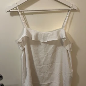 Vit volangprydd topp  från H&M - Säljer en vit tooo från H&M med smala axelband och volangdetalj längs överkanten. Toppen har en luftig och lätt känsla, perfekt för varma dagar. 