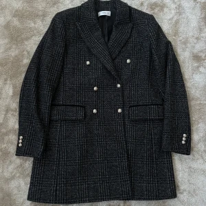 Svart rutig kappa från MNG - Säljer en svart och grå rutig kappa från MNG med dubbelknäppning och dekorativa silvriga knappar. Klassisk krage och två fickor framtill. Perfekt för dig som gillar en stilren och tidlös look.