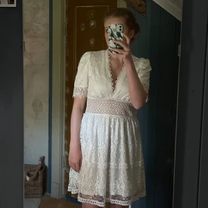 Vit spetsklänning med v-ringning - Säljer en vit kortklänning med v-ringning och fina spetsdetaljer. Klänningen har korta ärmar, markerad midja och vackert mönstrad spets över hela plagget. Perfekt för sommarens tillfällen eller student. Aldrig använd 