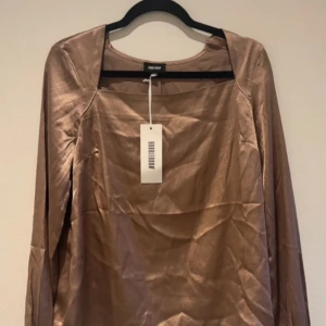 Glansig brun långärmad topp med fyrkantig ringning - Säljer en glansig  långärmad topp från bubbleroom med fyrkantig ringning. Toppen har en elegant och stilren look, perfekt att styla till olika outfits. Materialet är mjukt och har en satinliknande finish. Färgen är brun/lila