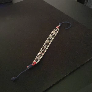 Svart och rött armband med mönster och nitar - Säljer ett handgjort armband med svart, vitt och rött broderat mönster. Armbandet har dekorativa guldfärgade nitar och knytband för justerbar passform. Perfekt accessoar för att ge din outfit en unik touch. Man kan justera den så den passar alla. De är bara att skriva ifall du har funderingar eller frågor 😁