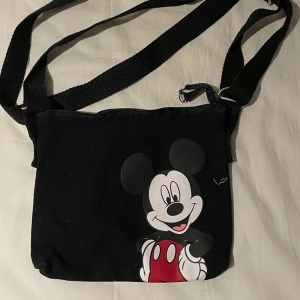 Svart axelväska med Musse Pigg-motiv - Säljer en svart axelväska med ett stort tryck av Musse Pigg på framsidan. Väskan har justerbar axelrem och stängs med dragkedja upptill. Perfekt för dig som gillar Disney och vill ha en rolig accessoar till vardagen.