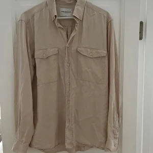 Beige skjorta från These Glory Days - Säljer en stilren beige skjorta från These Glory Days i mjukt lyocellmaterial. Skjortan har två bröstfickor med lock och knappar samt klassisk krage. Perfekt för en clean och avslappnad look.