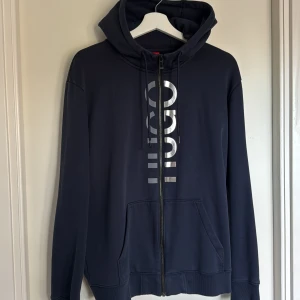 Mörkblå hoodie med dragkedja från Hugo - Säljer en mörkblå hoodie från Hugo med dragkedja och silverfärgat tryck framtill. Tröjan har huva med dragsko och två fickor framtill. Perfekt för en avslappnad stil.