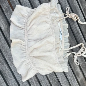 Beige volangtop från NAKD - Supersöt beige linnetopp från NAKD med volangdetaljer och smala axelband som knyts upptill. Tunt, luftigt material som passar perfekt till varma dagar. Toppen har en lätt rynkad struktur och en feminin, romantisk look.