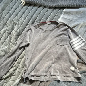 Grå långärmad tröja Thom Browne - Grå långärmad tröja från Thom Browne med klassiska vita ränder på ena ärmen och liten bröstficka. Tröjan har rund hals, knappar i sidan och diskret etikett nertill. Tillverkad i mjuk bomull och har röd, vit och blå detalj i nederkant.