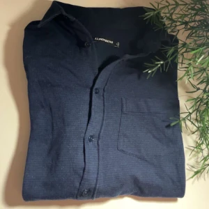 Mörkblå skjorta från J.Lindeberg - Snygg mörkblå skjorta från J.Lindeberg med klassisk krage, bröstficka och knäppning framtill. Skjortan är långärmad och har en diskret textur i tyget som ger en cool känsla. Perfekt för dig som gillar stilrena plagg med en modern touch.