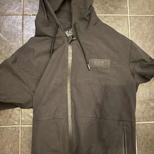 Svart vindjacka från EA7 Emporio Armani - Svart vindjacka från EA7 Emporio Armani med huva och hel dragkedja framtill. Jackan har diskret logga på bröstet, dragsko i huvan och två fickor med dragkedja. Perfekt för en clean och sportig look.