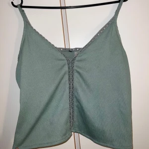 Grön ribbad linnetopp med spetsdetalj - Snygg ljusgrön linnetopp med smala axelband och v-ringning. Toppen har ribbad struktur och fina spetsdetaljer längs urringningen och framtill. Perfekt för varma dagar och enkel att matcha med jeans eller kjol.