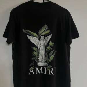 Svart Amiri t-shirt med ängeltryck - Svart t-shirt från Amiri med stor grafisk ängel och gröna blad på ryggen samt Amiri-logga. Framsidan har tryckt logga med färgstänk. Klassisk rund hals och korta ärmar. Skön passform och riktigt snygg streetstil.