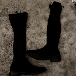 Svarta höga mockastövlar med dragkedja - Snygga svarta overknee-stövlar i mocka med dragkedja på insidan och låg klack. Skorna har en klassisk rund tå och platt sula, perfekta för att lyfta din outfit under kallare månader.