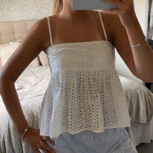 Vit broderad linnetopp med spets - Söt vit linnetopp med tunna axelband och broderade hål för en luftig känsla. Toppen har en lätt utsvängd passform och spetsdetalj längs överkanten. Perfekt för varma dagar och ger en romantisk vibe.