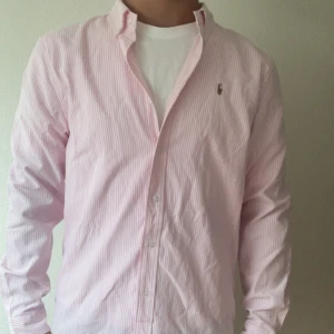 Rosa randig skjorta från Polo Ralph Lauren - Säljer en klassisk rosa och vit randig skjorta från Polo Ralph Lauren. Skjortan har lång ärm, knappar framtill och broderad logga på bröstet. Classic fit-modell i mjuk bomull, perfekt till jeans eller chinos.