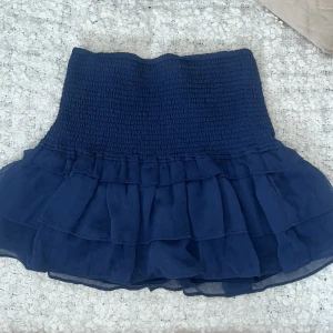 Mörkblå volangkjol med smock - Supersöt mörkblå kortkjol med bred smock i midjan och flera lager volanger. Kjolen har en luftig och flowy känsla, perfekt för att skapa en trendig och avslappnad look. Passar lika bra till sneakers som boots.